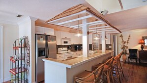 Une cuisine moderne avec des meubles blancs, un îlot central doté de tabourets de bar et un plafond au style rustique.