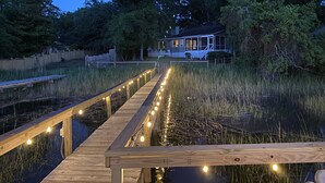 A wooden deck with string lights leading to a house surrounded by trees and a fence. - Shem Creek Paradise. Fish, Paddle, Kayak Off Your Own Covered dock with floater (Mt Pleasant)
