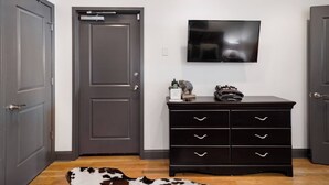 A room with a dark wooden dresser, a flat-screen TV on the wall, and a black and white dog lying on the floor. - On Broadway Above Boot Barn (Nashville)