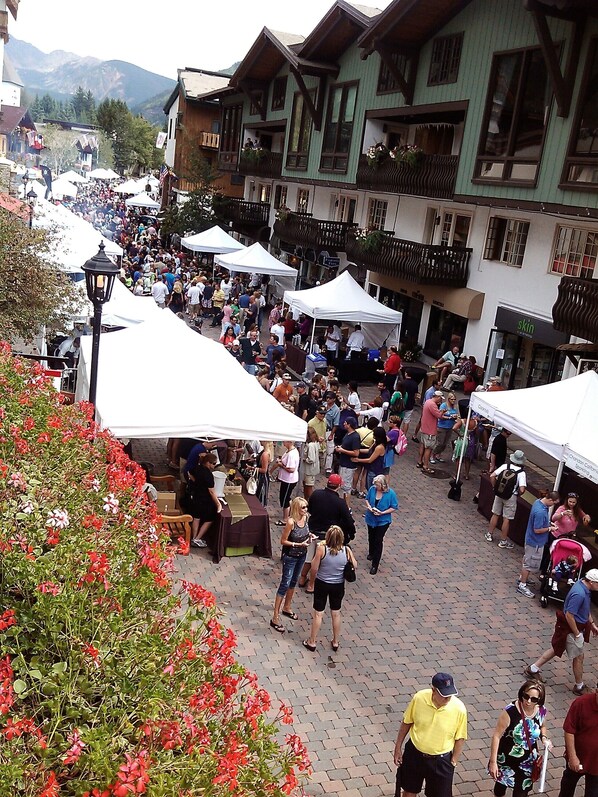 A bustling outdoor market with people walking and vendors under tents, set against a backdrop of traditional buildings and mountains.