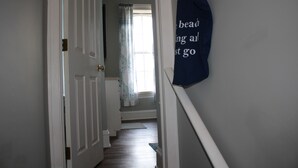 Un pasillo con una puerta blanca, una ventana con cortinas y un bolso azul colgado en la pared.