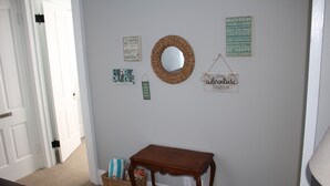Un pasillo con una mesa de madera, una cesta de mimbre y varias decoraciones en las paredes, incluyendo un espejo, letreros enmarcados y un letrero colgante.
