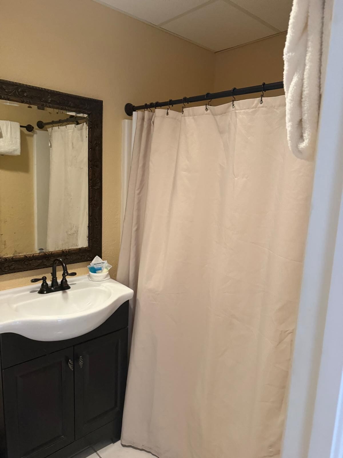 A bathroom with a white sink, dark cabinets, a mirror with an ornate frame, and a beige shower curtain.
