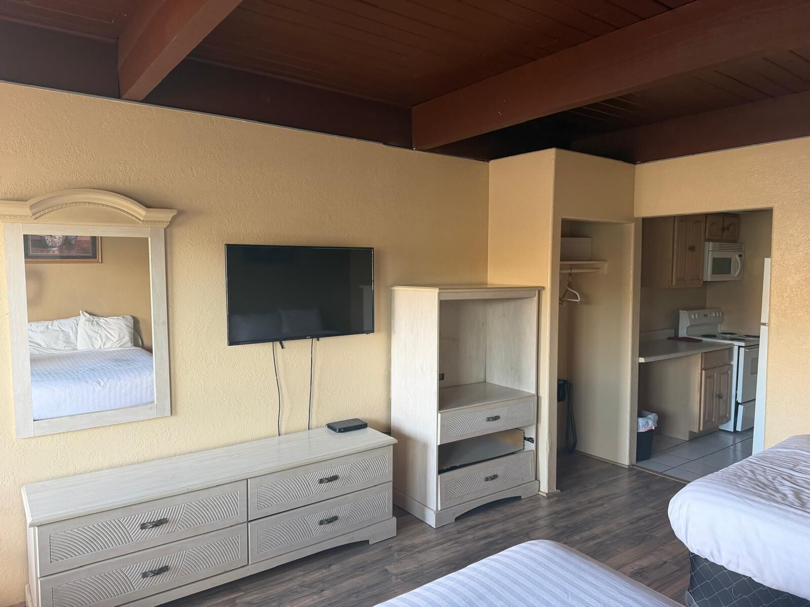 A hotel room with a bed, a TV mounted on the wall, a dresser, and a kitchenette area.
