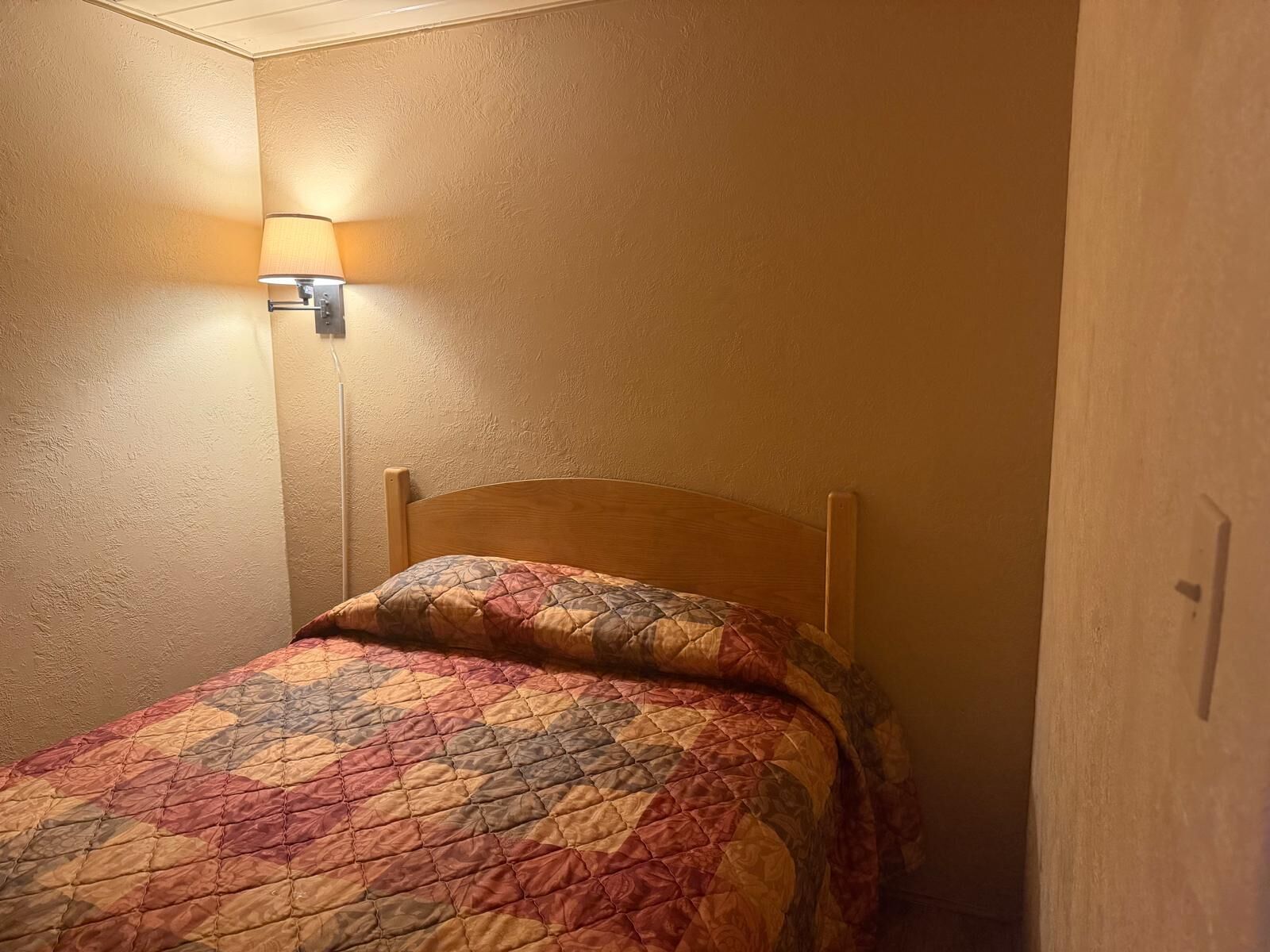 A single bed with a wooden headboard in a room with beige walls and a wall-mounted light fixture.