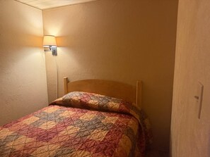 Una cama individual con cabecera de madera en una habitación con paredes color beige y una lámpara fijada a la pared.