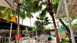 An outdoor seating area with white plastic chairs and tables, surrounded by palm trees and greenery. - Riviera Punta Cana Eco Travelers House (Punta Cana)