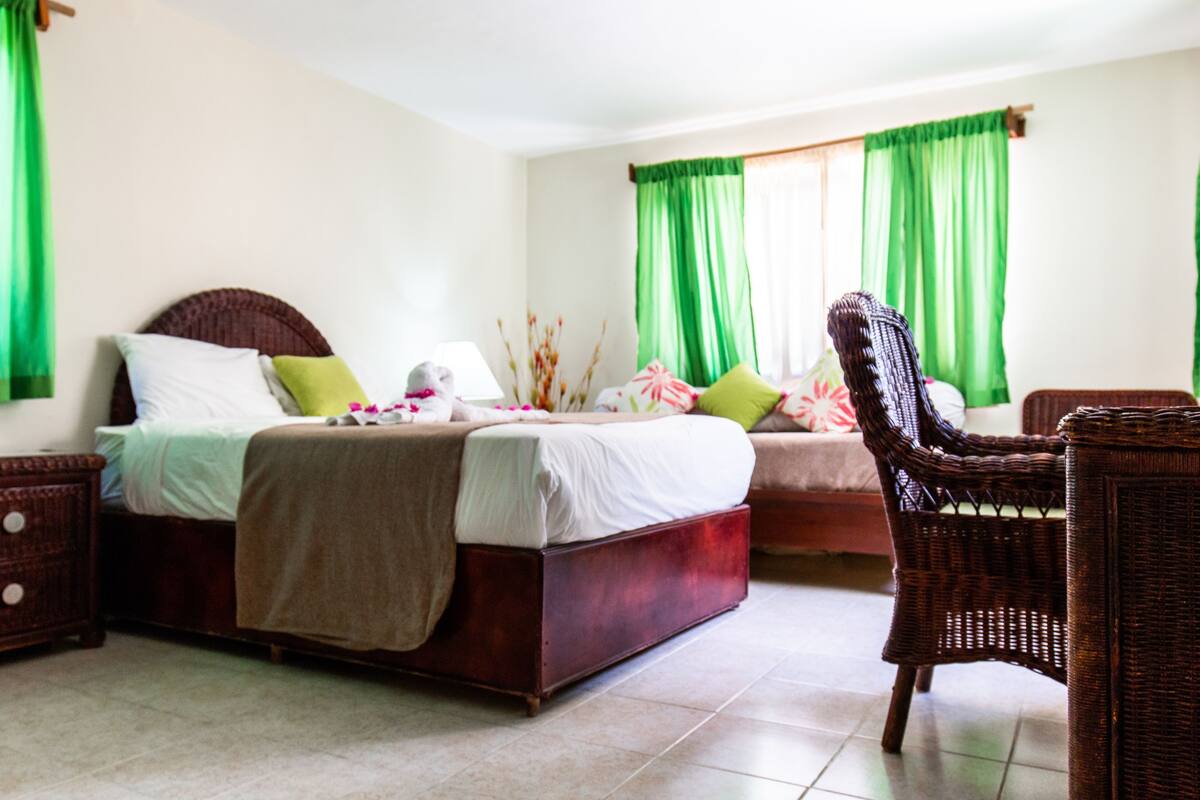 A bedroom with two beds, green curtains, a wicker chair, and a nightstand.