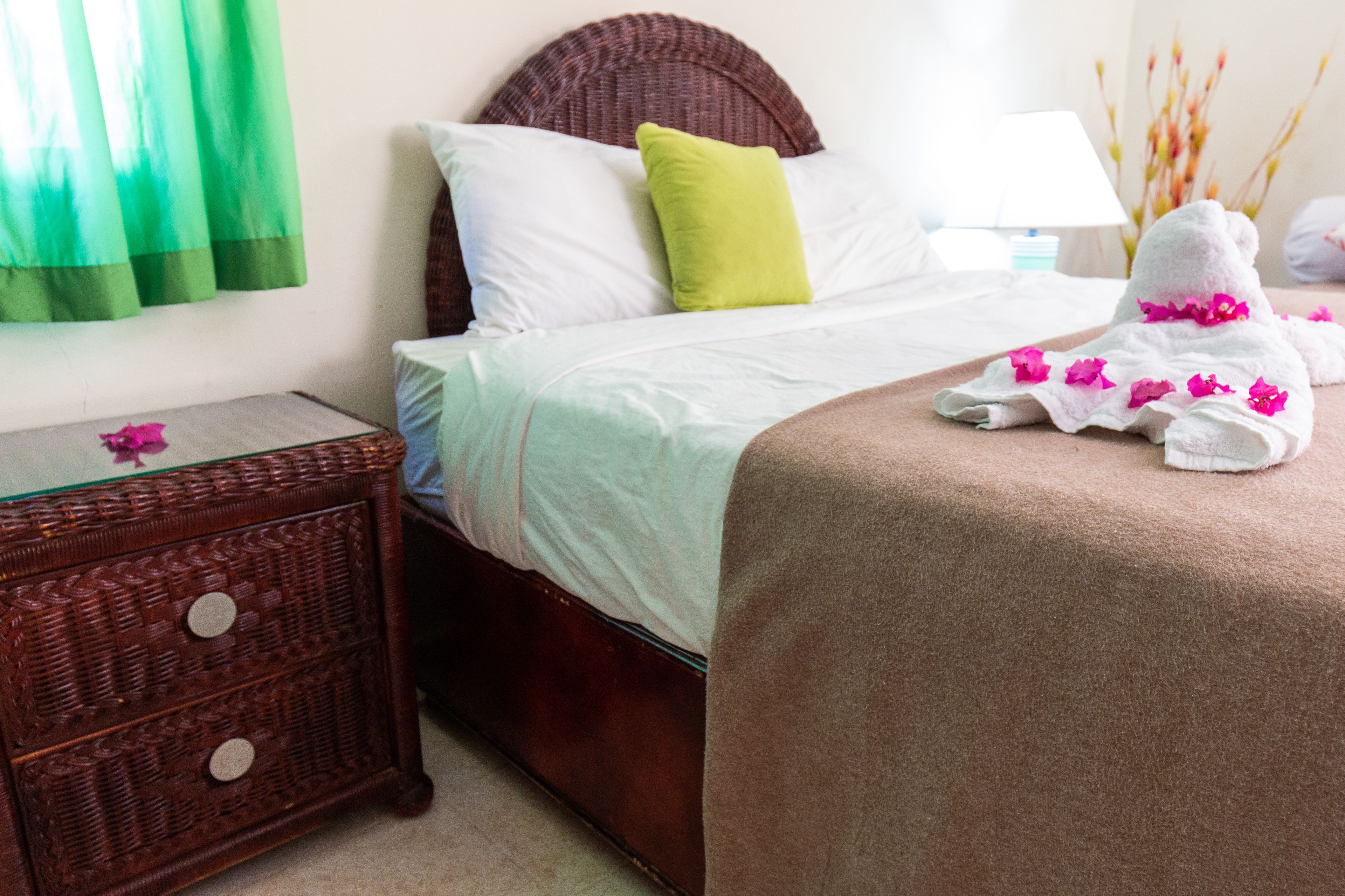 A neatly made bed with a green pillow, a wicker nightstand with a glass top, and a towel with pink flowers on a bed.