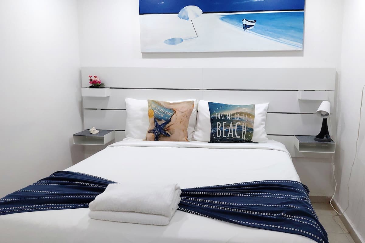 A neatly made bed with a white comforter and a blue and white striped blanket. There are two pillows with beach-themed designs. A painting of a beach scene is on the wall above the bed.