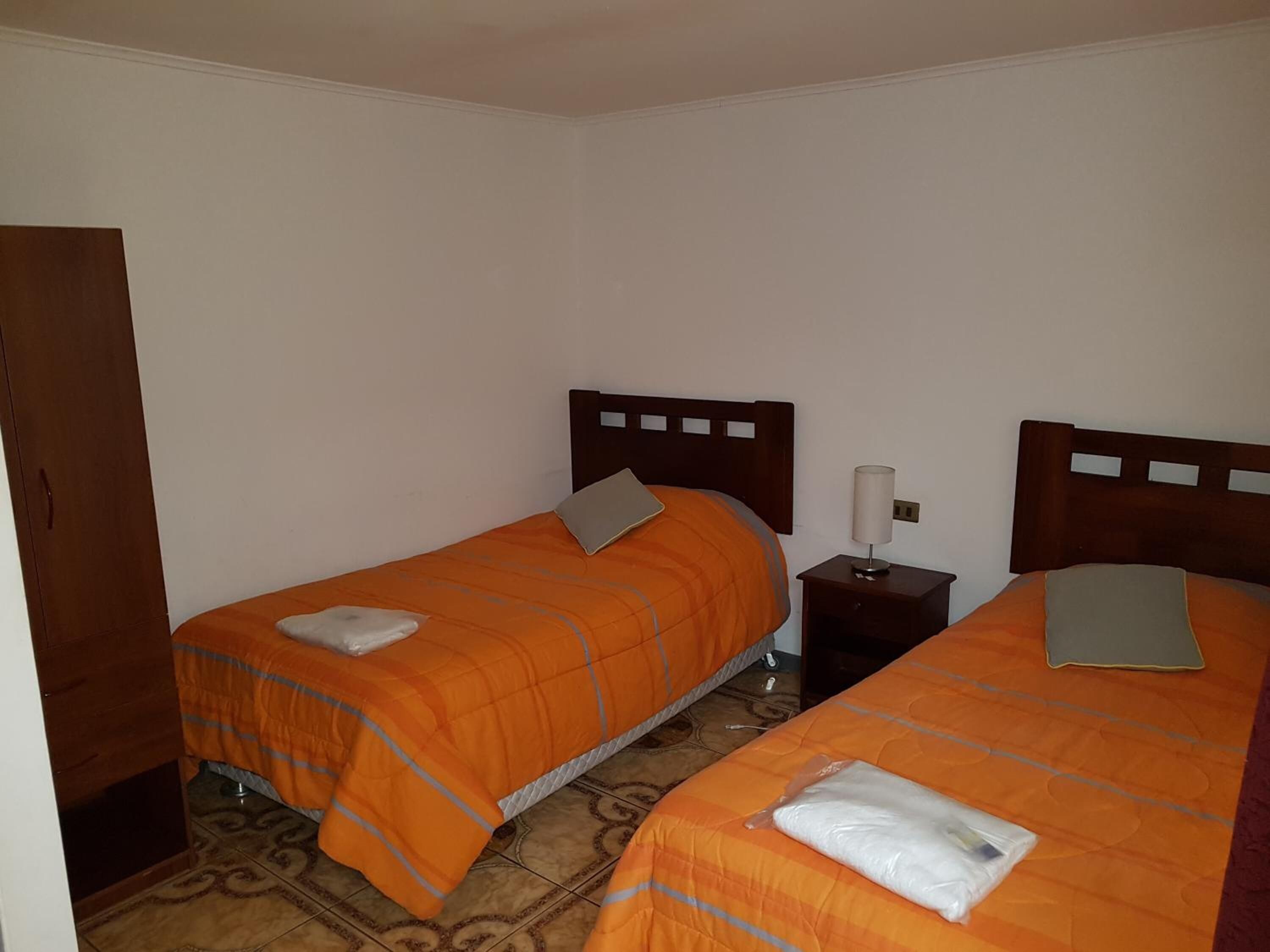 A room with two single beds, each with an orange bedspread and a pillow, a bedside table with a lamp, and a wooden wardrobe.
