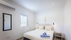 Une chambre moderne avec un grand lit, une table de chevet, une petite commode, une fenêtre et un appareil de climatisation.