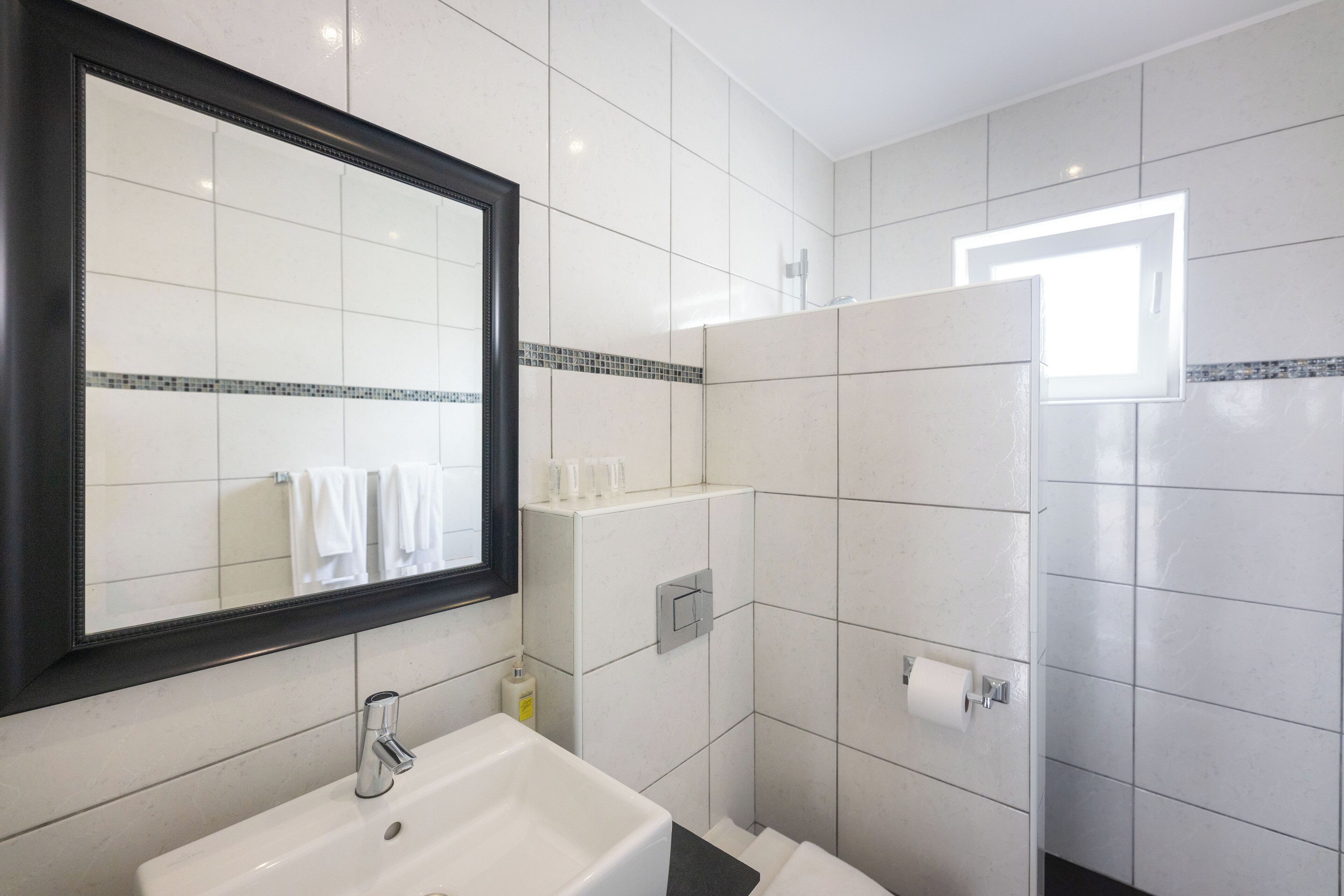 Un bagno moderno con un ampio specchio, piastrelle bianche, un lavandino e una zona doccia.