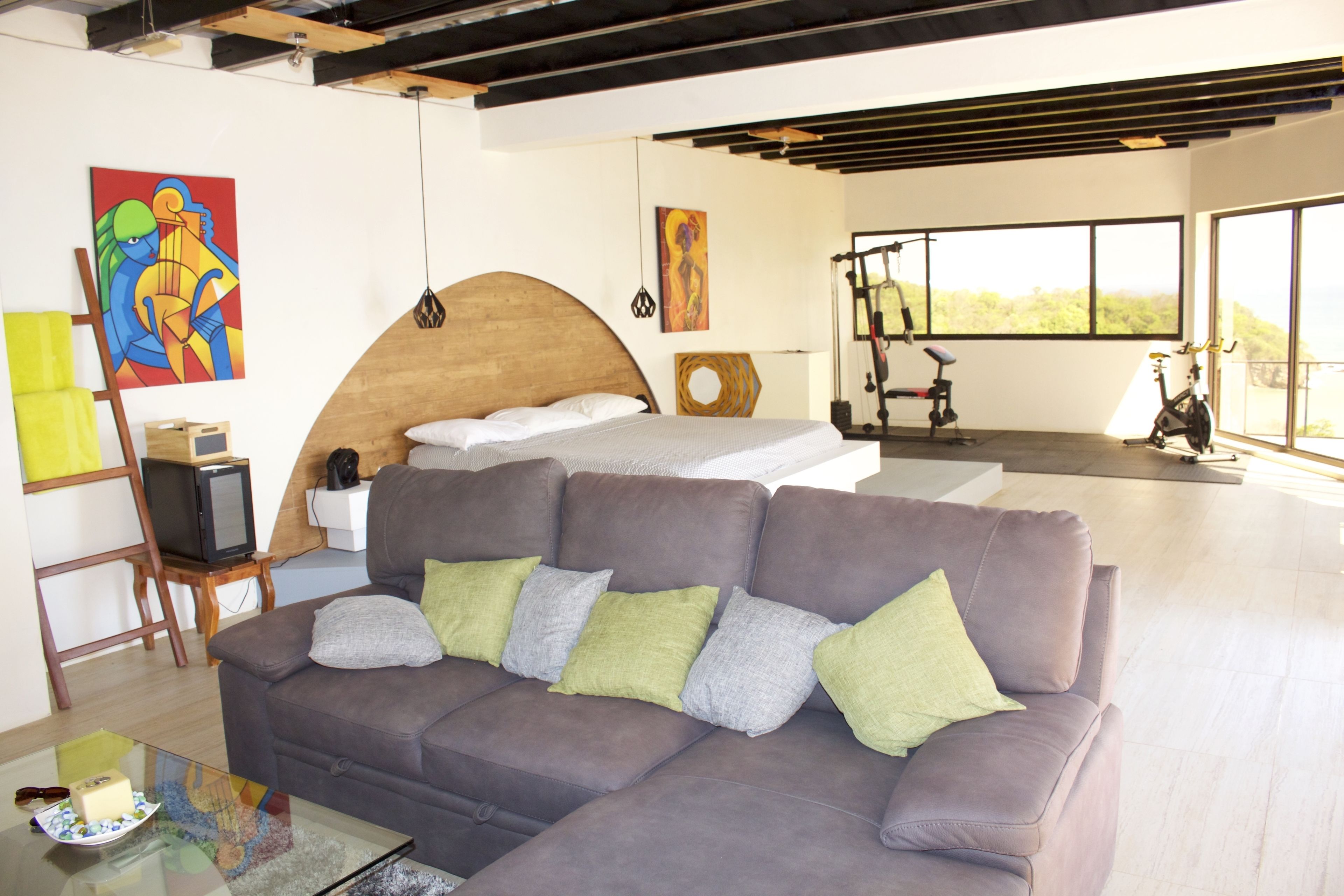 A modern living room with a large bed, a sofa, and a TV. There are paintings on the wall and a window with a view.