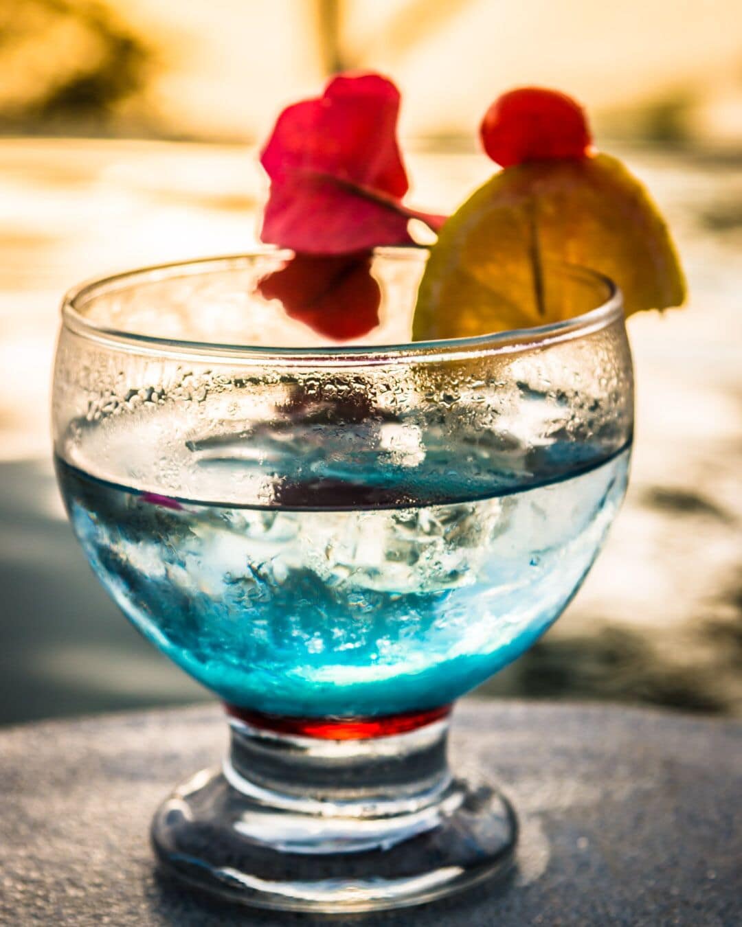 Un verre contenant un liquide bleu, peut-être un cocktail, agrémenté d’une fleur rouge et d’une tranche de citron vert.