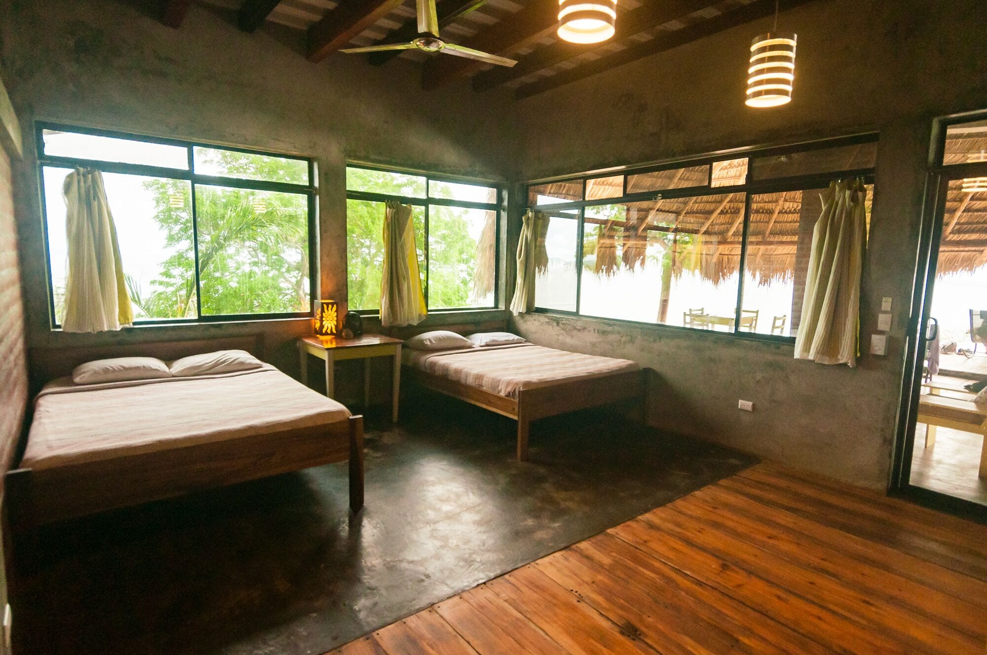 A room with two beds, a small table with a lamp, large windows, and a ceiling fan.