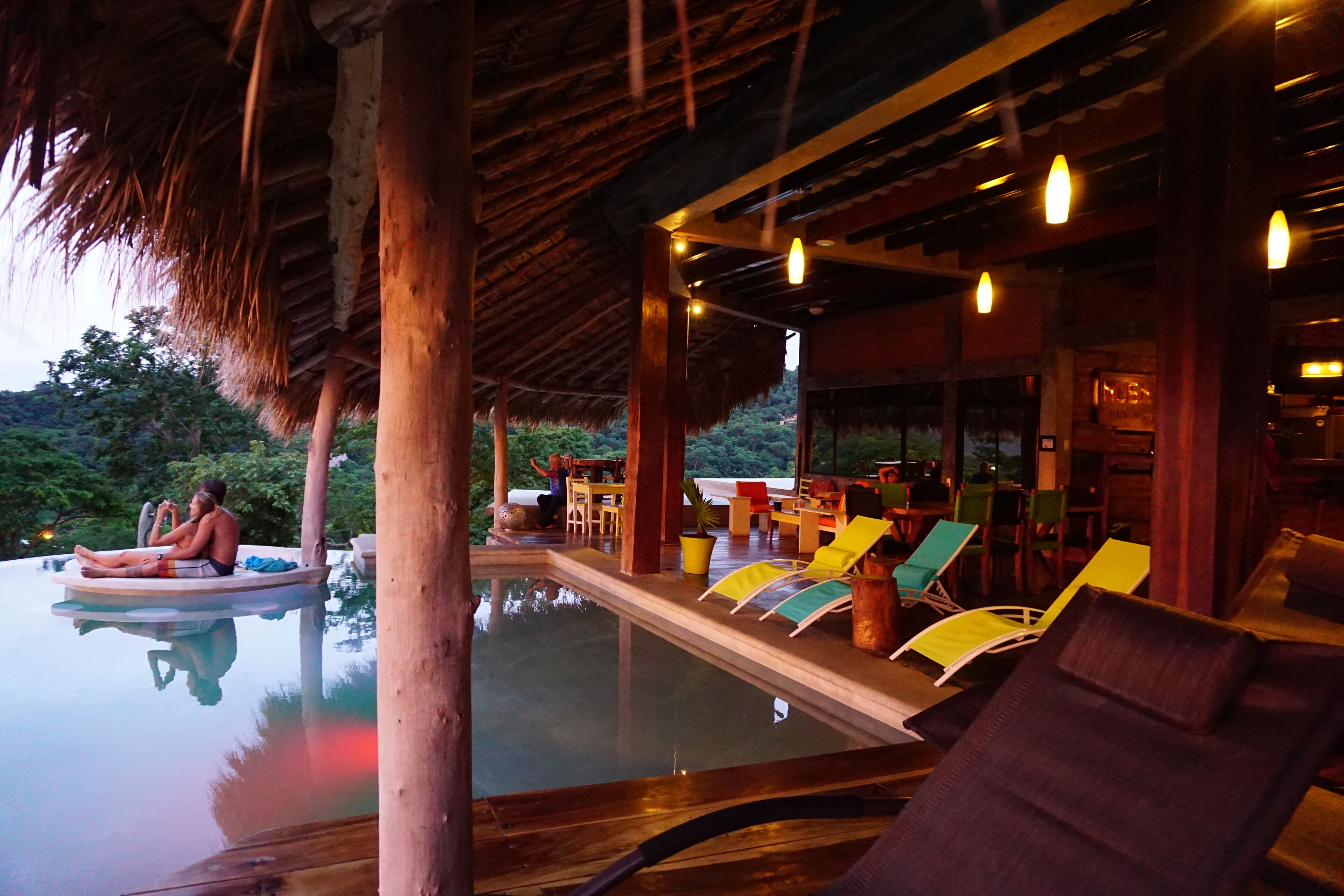 Un área junto a la piscina con sillones de descanso, una terraza de madera y una estructura de techo de paja.