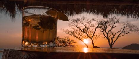 A glass of amber liquid with a lemon slice on a wooden surface, set against a sunset with silhouetted trees and a thatched roof structure.