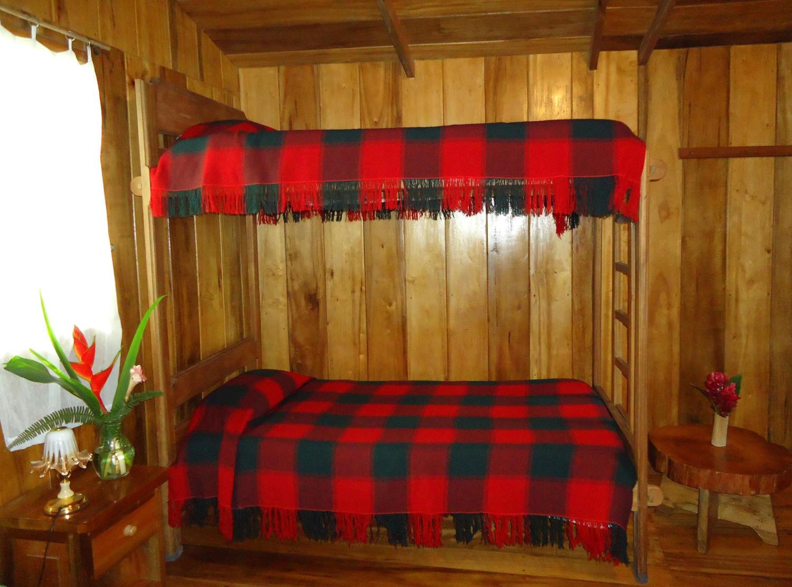 Une pièce de cabane en bois avec un lit recouvert d’une couverture à carreaux rouges et noirs, une petite table en bois avec une lampe et un vase rempli de fleurs.