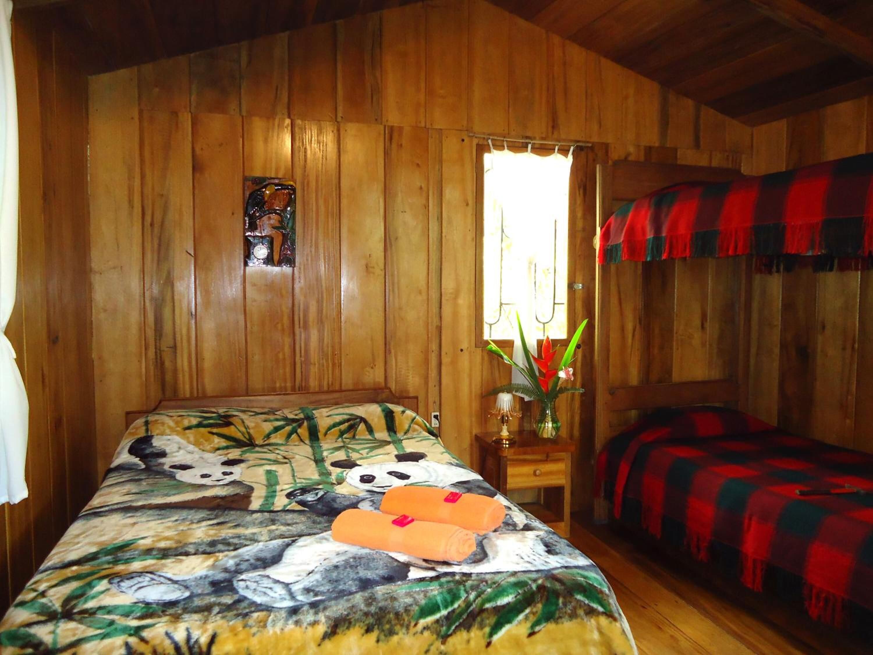 A wooden cabin room with a bed featuring a panda print, a small table with a vase of flowers, and a window with curtains.