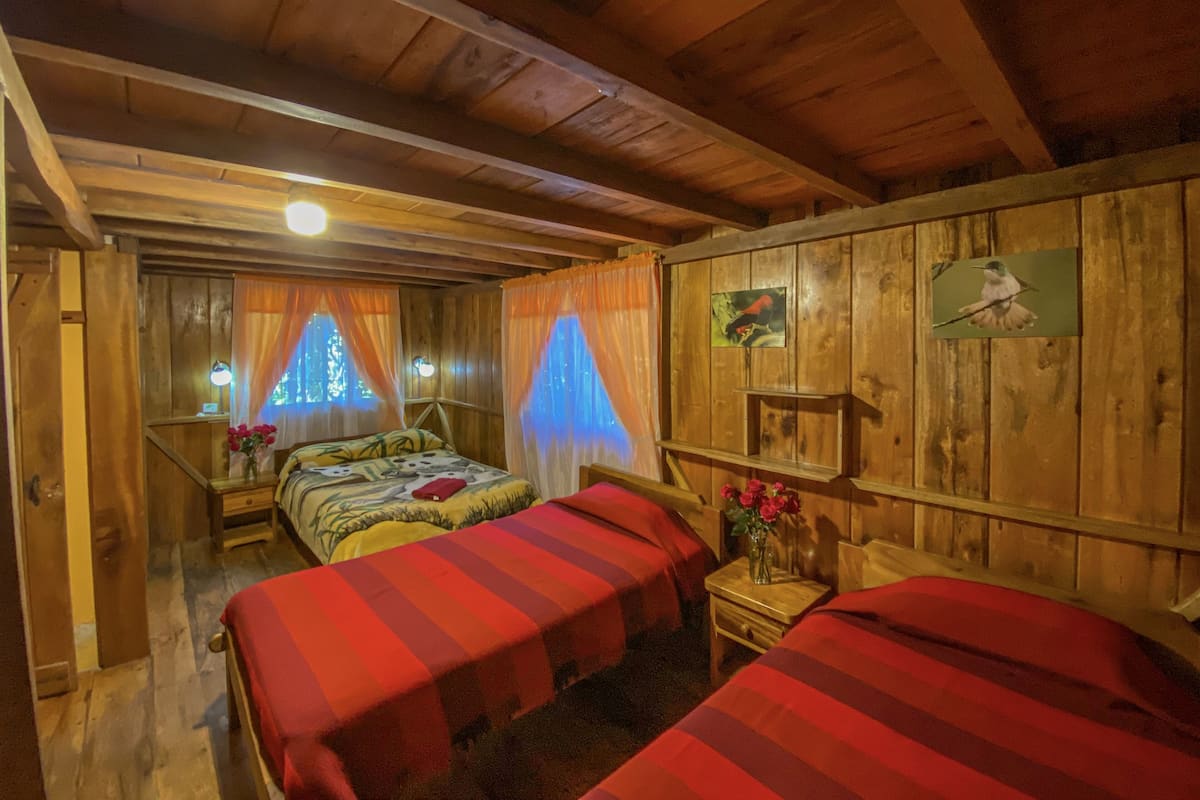 A wooden cabin room with two beds, a small table, and a window with curtains.