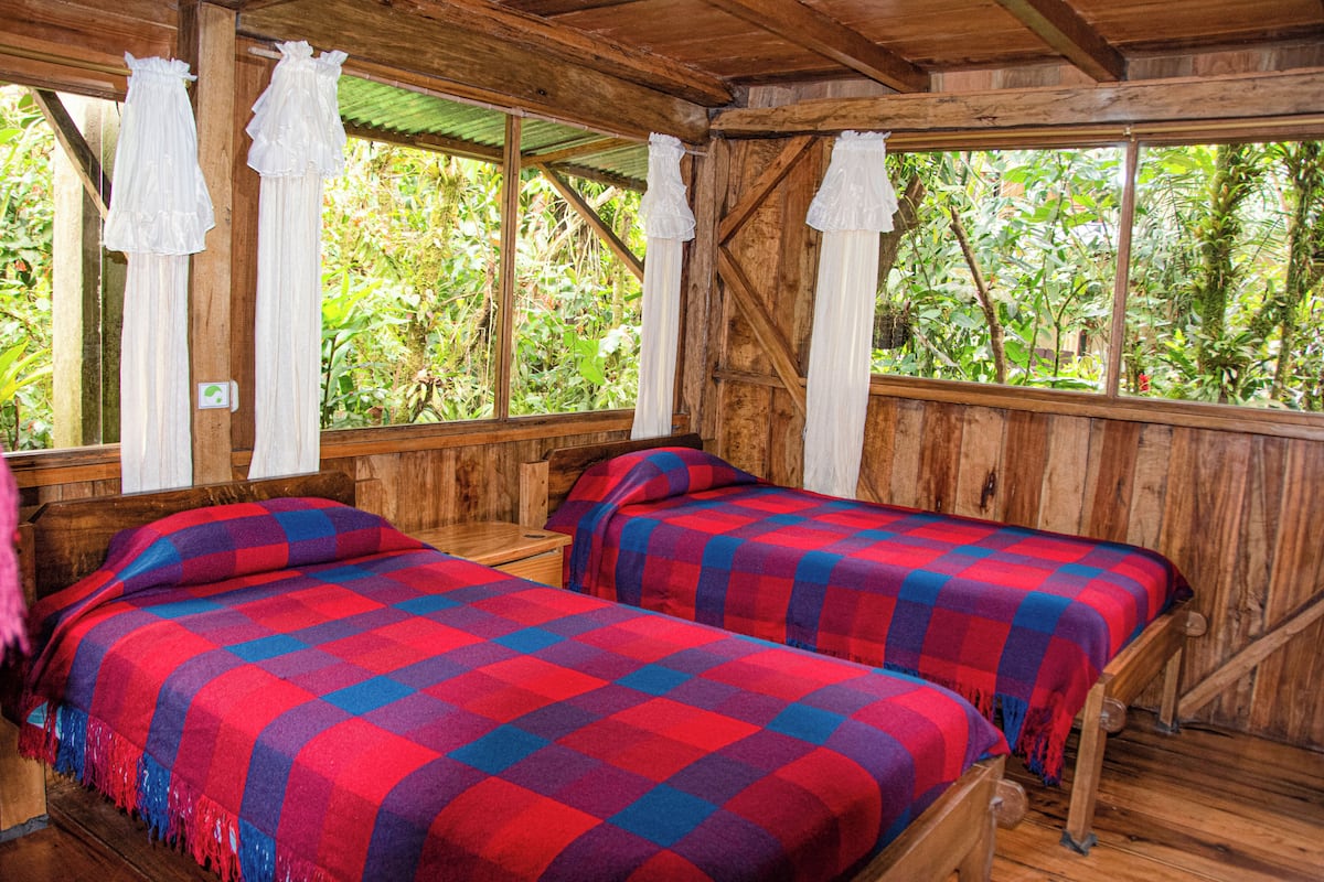 A wooden cabin with two beds, each with a red and blue checkered blanket, and white curtains.
