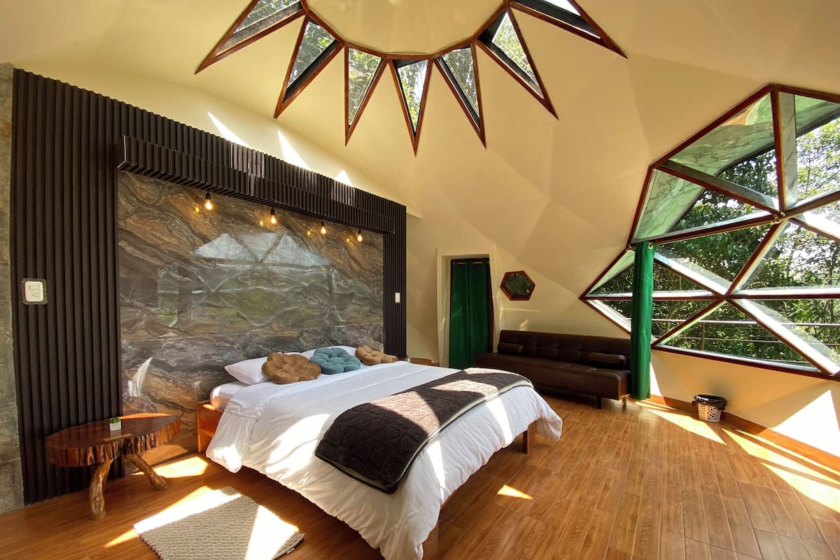 A bedroom with a large bed, a wooden floor, a stone wall, and a window with a geometric design.
