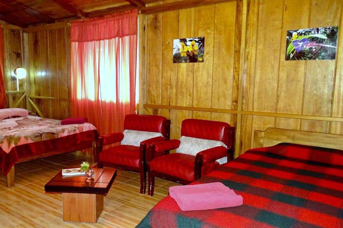 A wooden room with two beds, two red leather chairs, a small table, and a red and black checkered blanket.