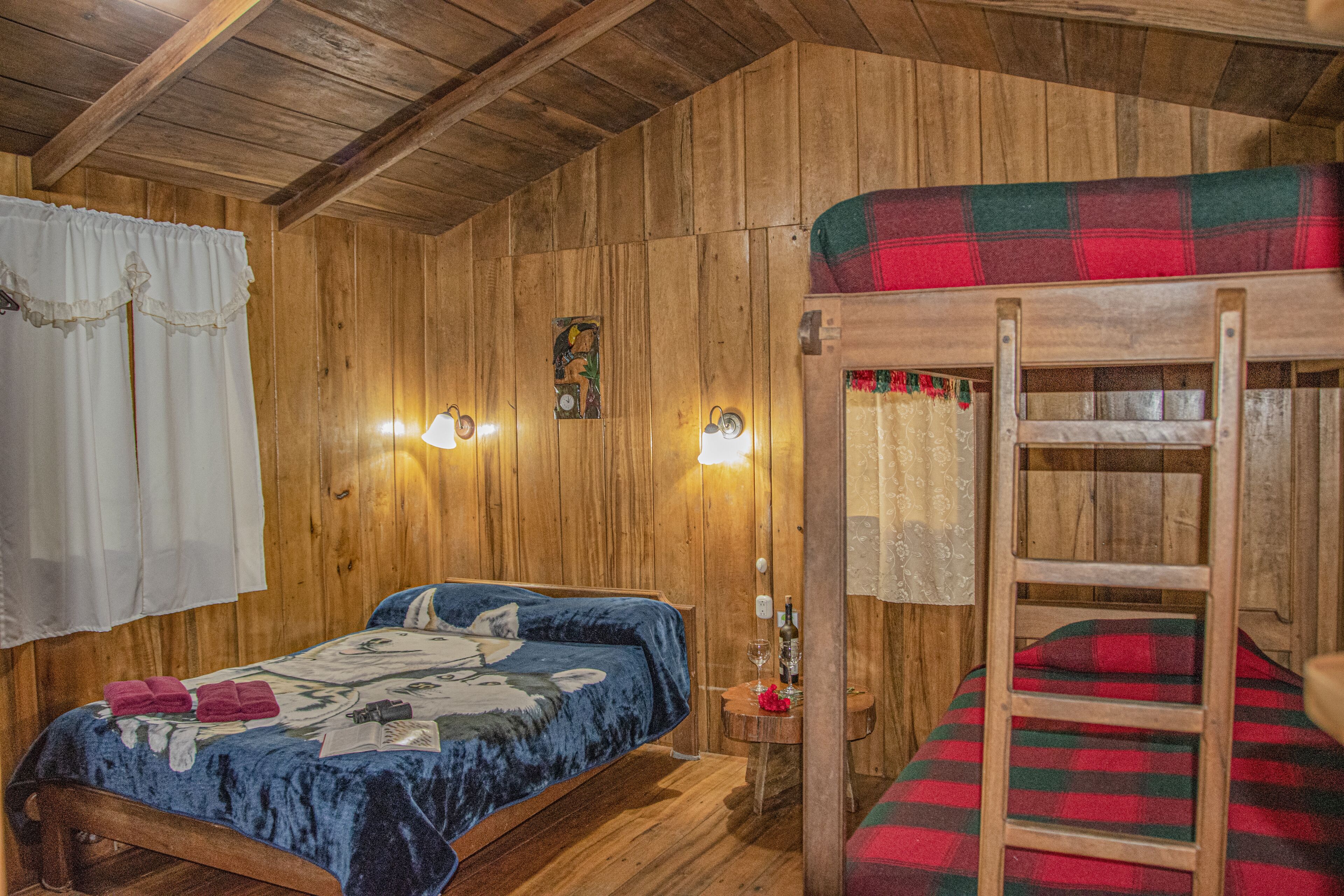 Ein Holzhüttenraum mit einem Stockbett, einem Einzelbett und einem kleinen Tisch mit einer Flasche und Gläsern.