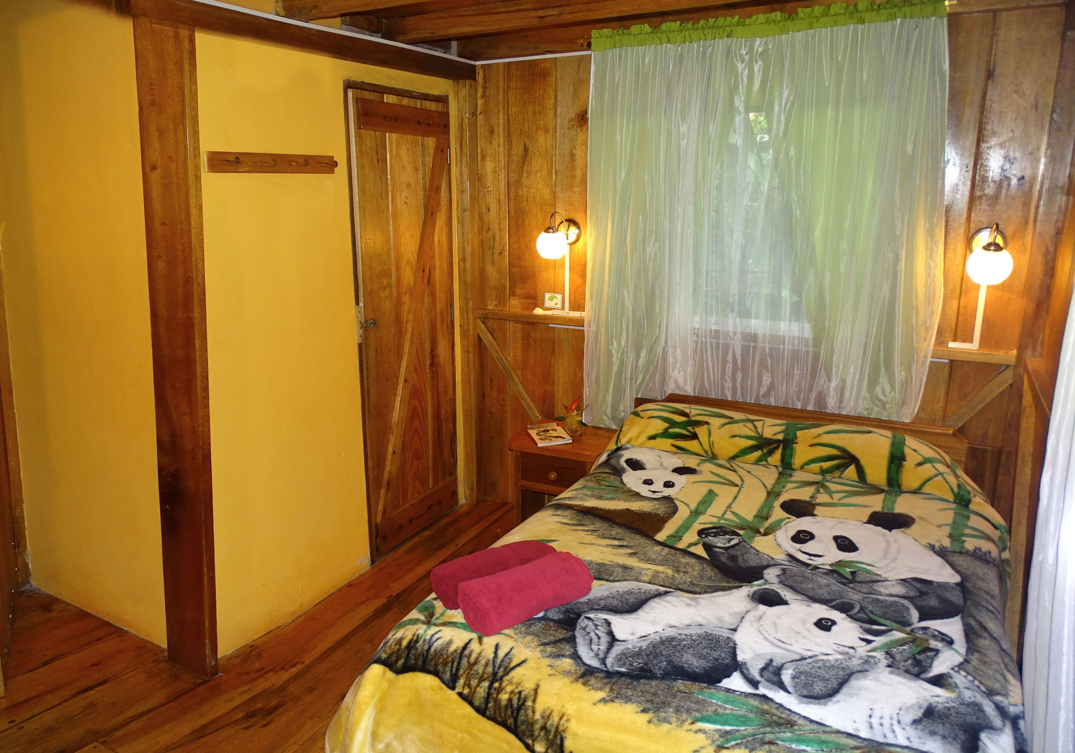 Een kamer met houten wanden en vloeren, een bed met een beddegoed met pandabeeld, twee bedlampen en een groen gordijn.