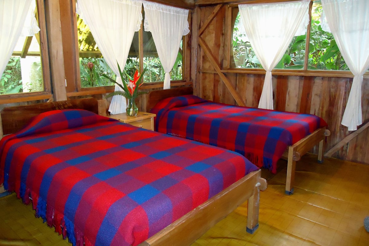 A room with two wooden beds, each with a red and blue checkered bedspread. There is a wooden nightstand with a vase of flowers between the beds. The room has wooden walls and windows with white curtains.