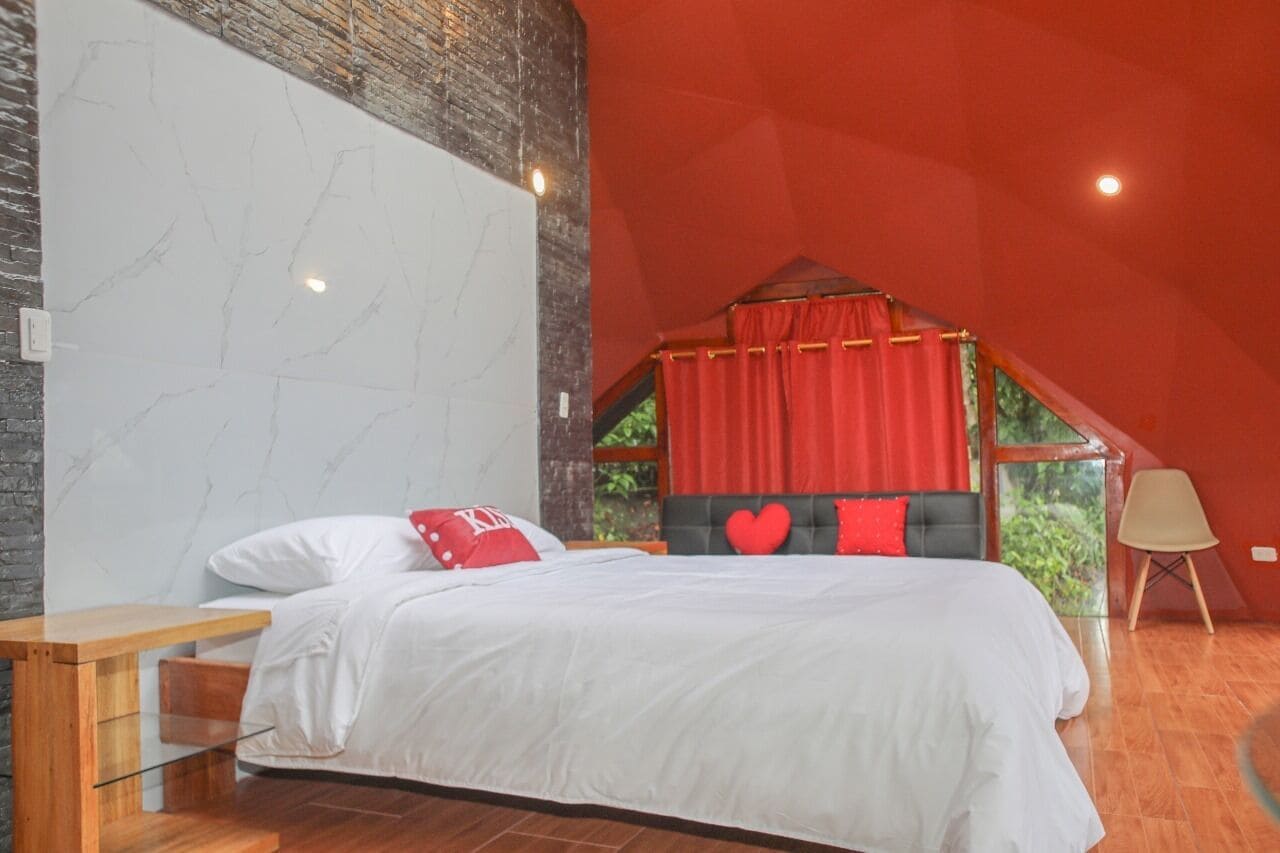 Ein Schlafzimmer mit einem großen Bett, einem hölzernen Nachttisch, einem roten Baldachinbett und Blick ins Grüne.