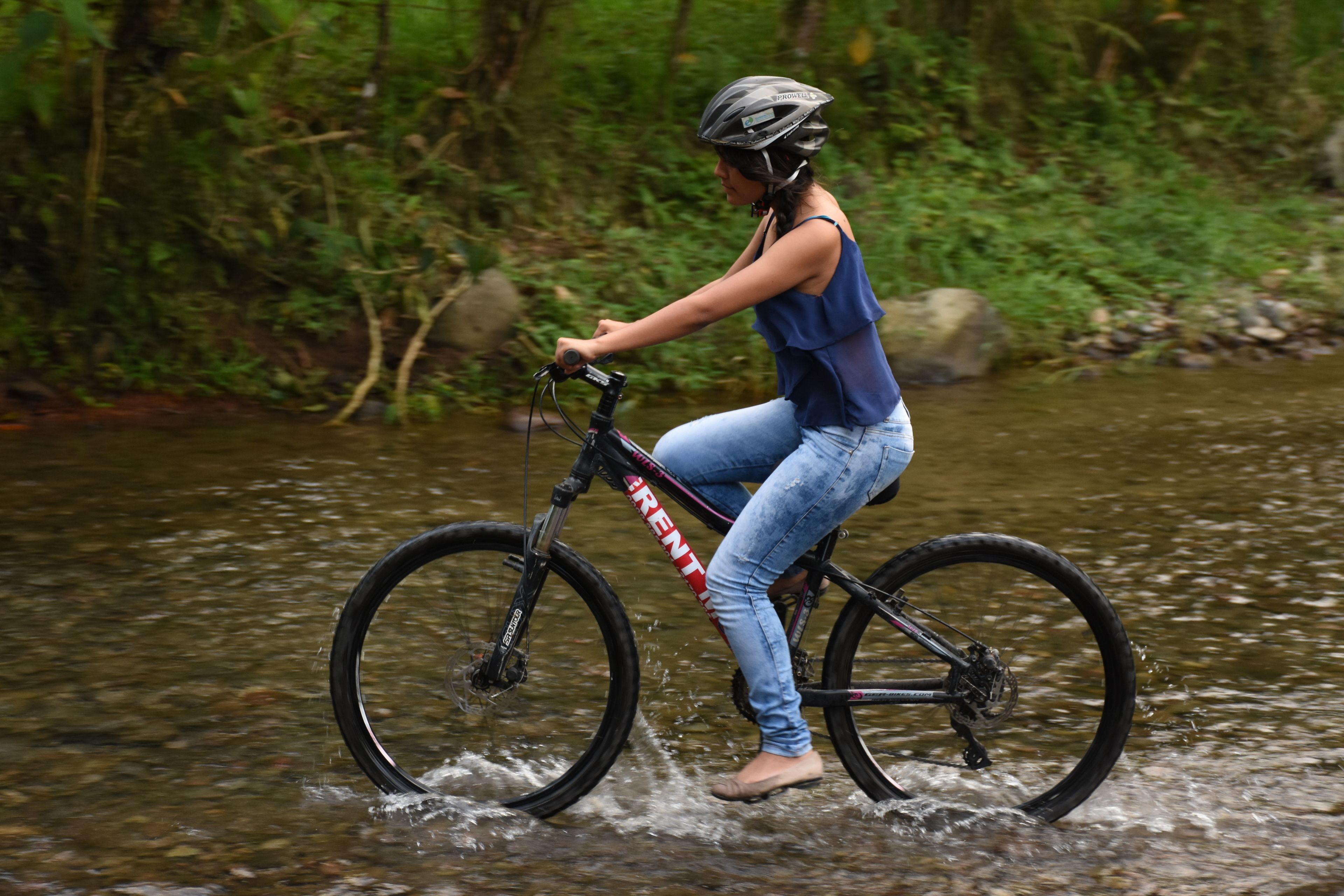 Una persona pedaleando una bicicleta por un arroyo poco profundo en un área boscosa.