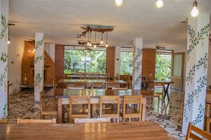 A spacious dining area with wooden tables and chairs, large windows with a view of greenery, and hanging light fixtures.