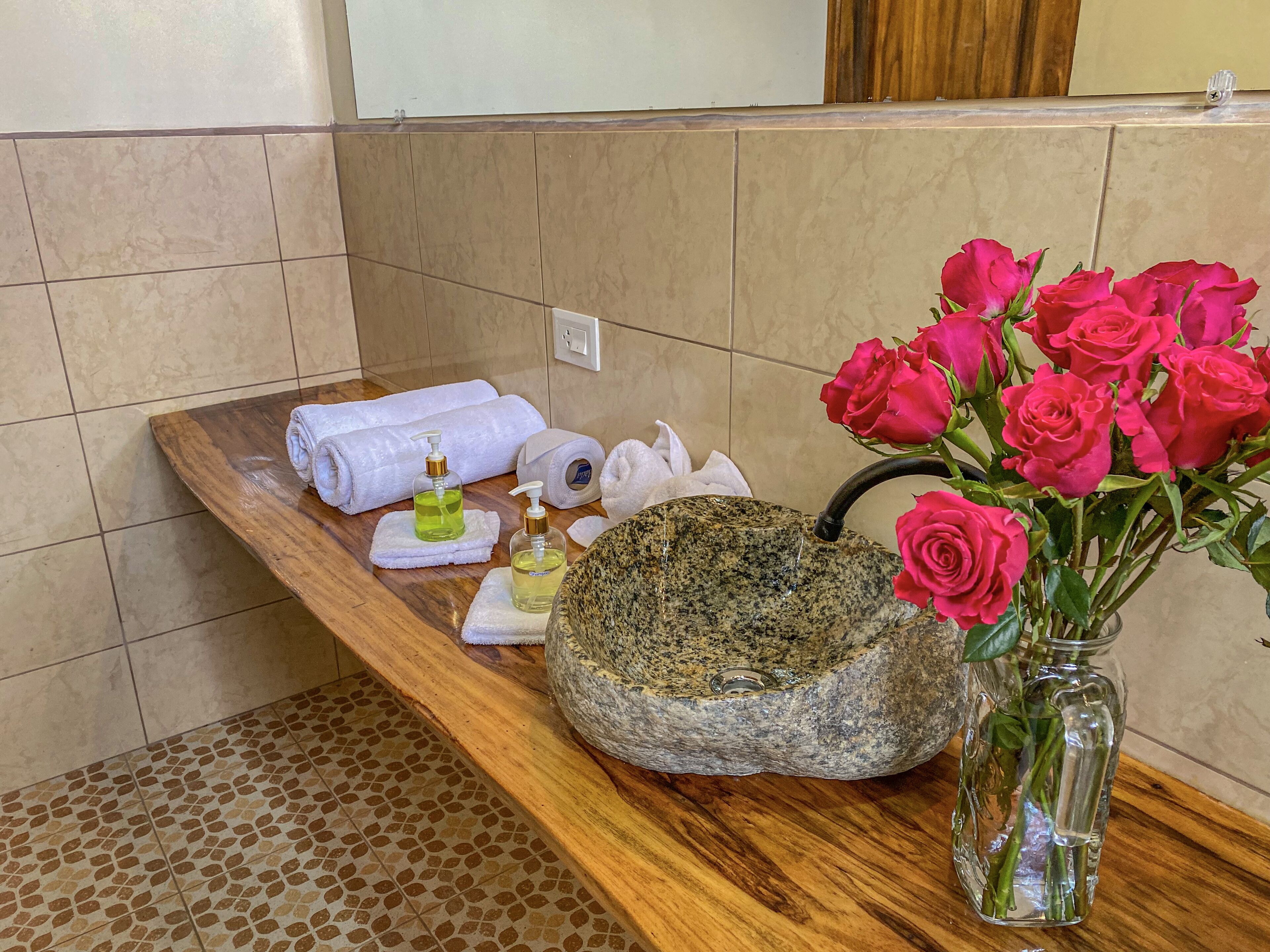 Un tocador de baño con un lavabo de piedra, un florero de rosas, artículos de aseo y toallas.