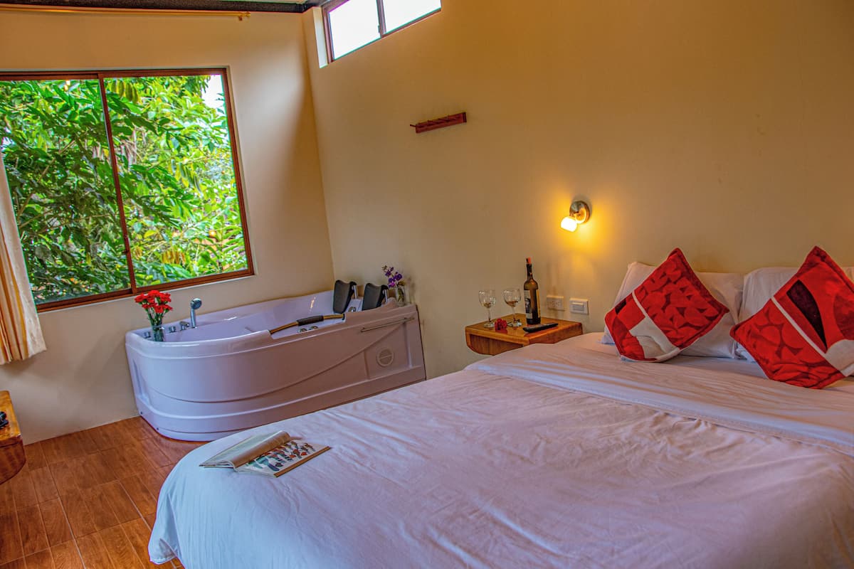 A hotel room with a large bed, a bathtub, a window with a view of greenery, a nightstand with a lamp, and a small table with a bottle and glasses.