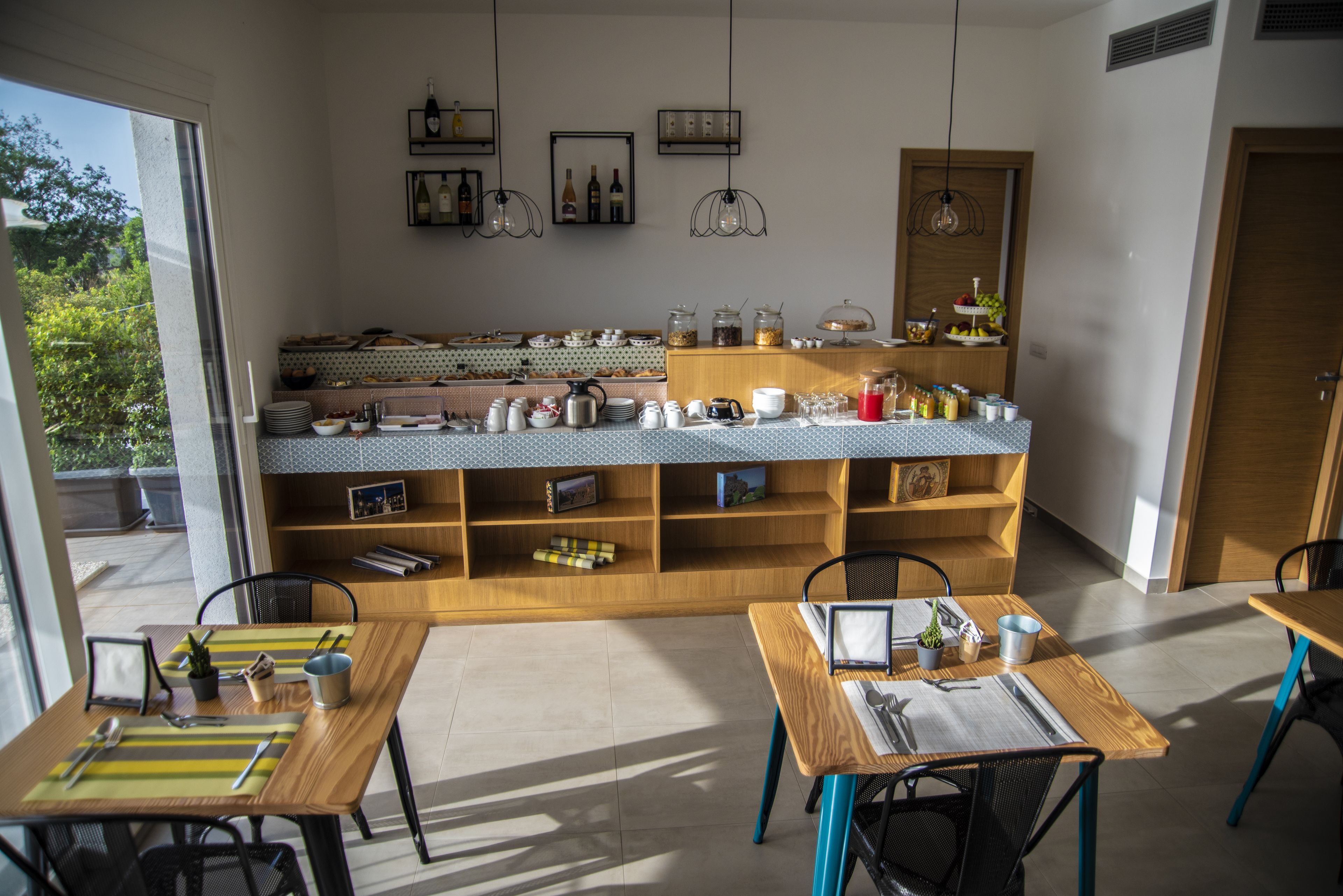 Un comptoir de petit-déjeuner proposant divers aliments et boissons, des tables en bois avec des chaises, et une vue sur la verdure environnante.