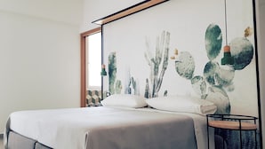 Un dormitorio con cama, almohadas, mesita de noche, un cuadro con cactus y una ventana con vistas.