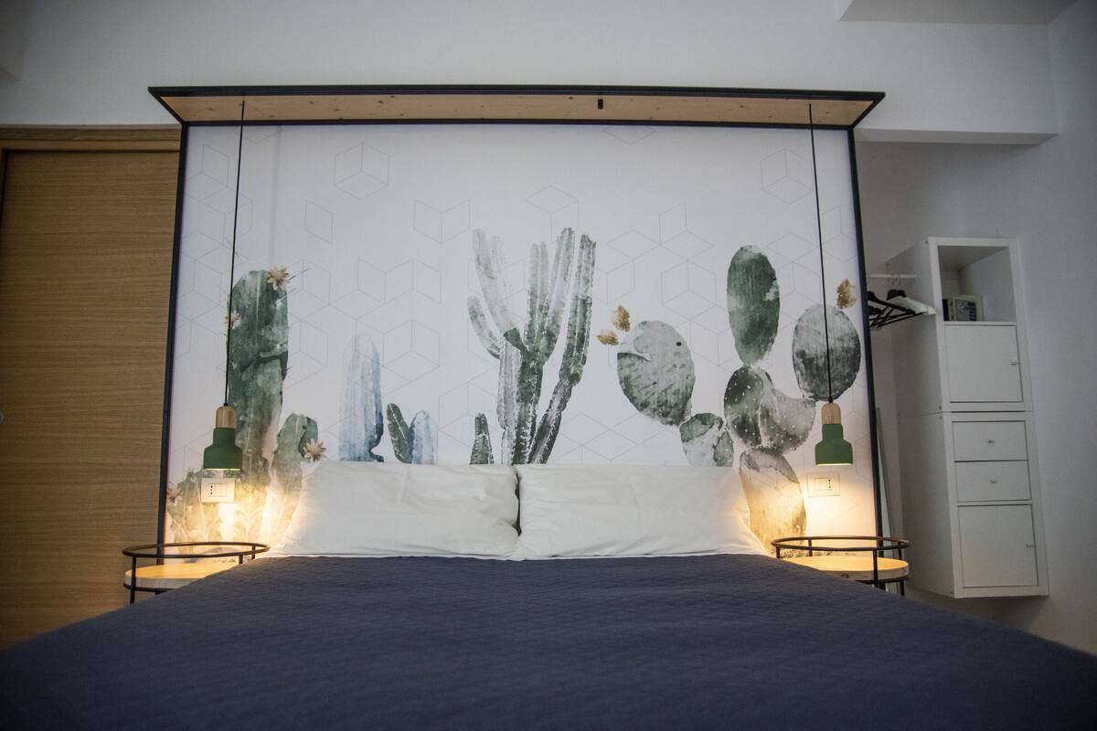 Un dormitorio con cama, dos mesitas de noche con lámparas, un armario blanco y un gran mural de cactus en la pared.