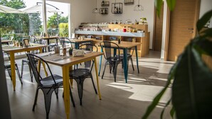 Un café moderno con varias mesas y sillas, una zona de cocina y ventanas amplias que dejan entrar luz natural.