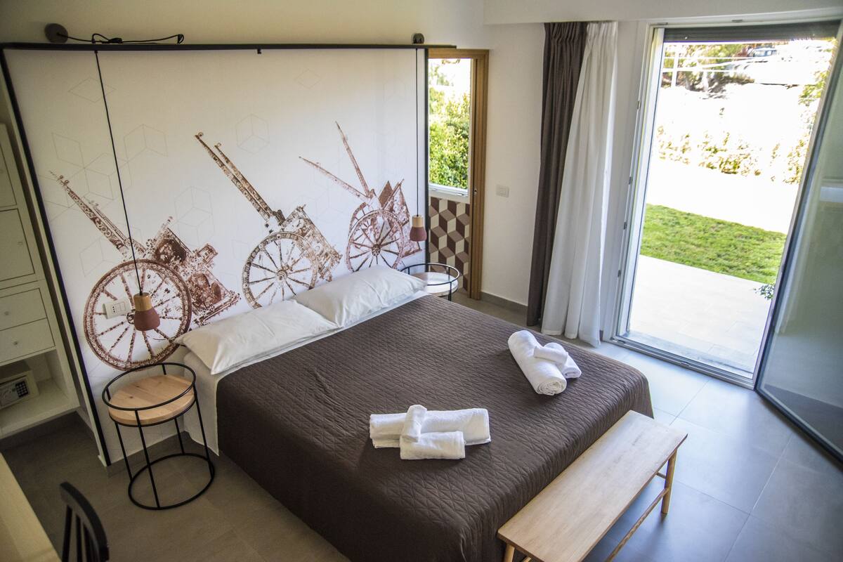 Un dormitorio con una cama grande, una mesita de noche, un taburete redondo y vistas al exterior a través de una puerta corredera.