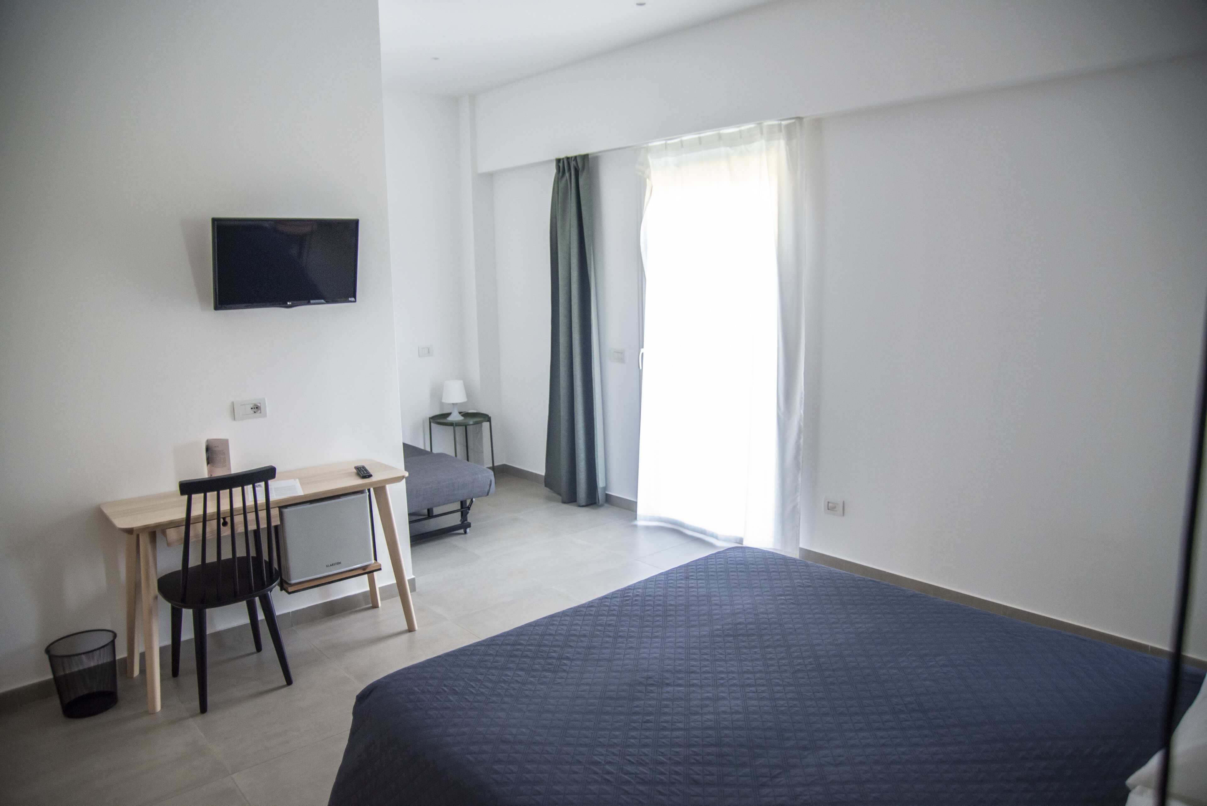 A bedroom with a bed, a desk, a chair, a TV, and a window with curtains.