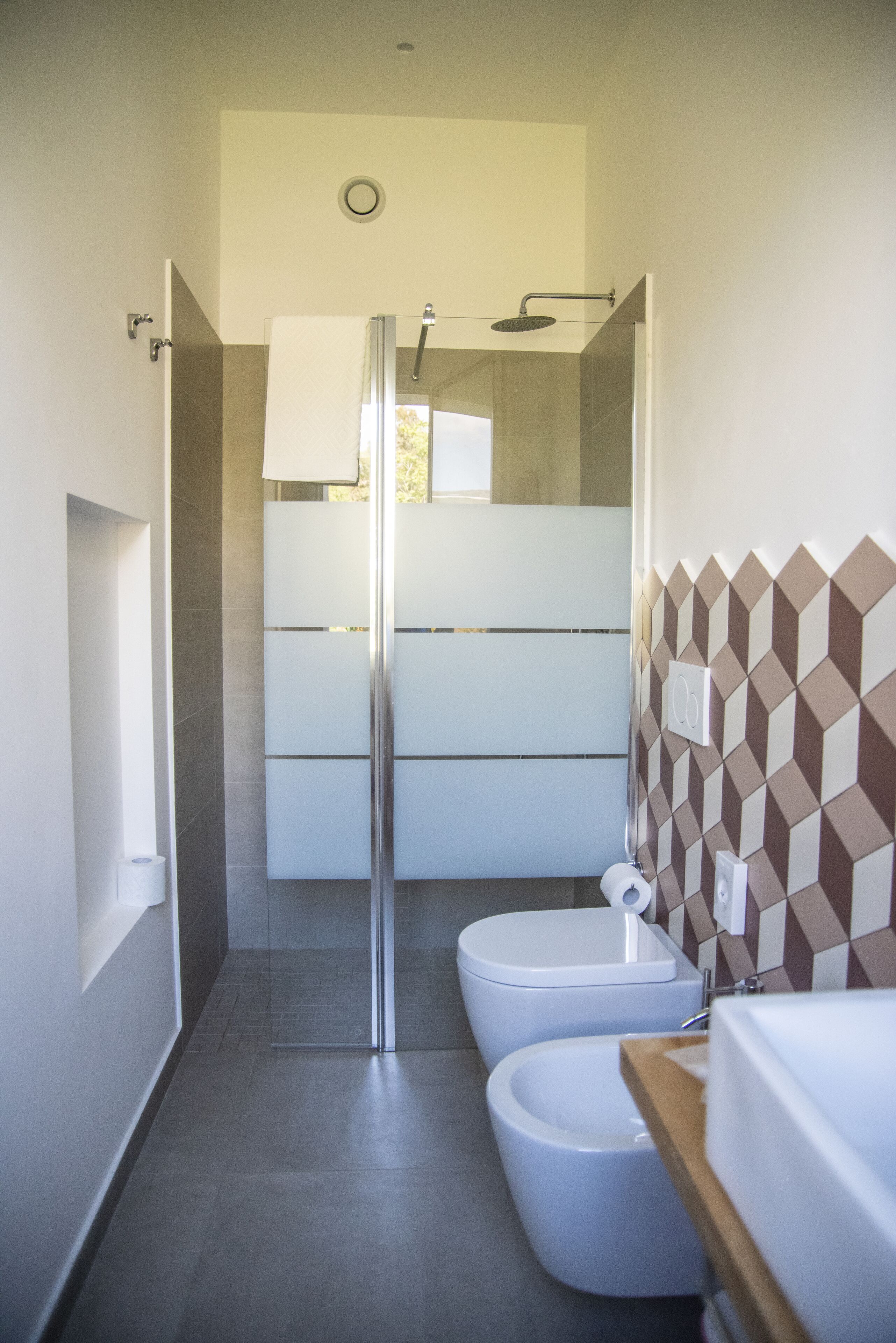 A modern bathroom with a shower, toilet, and bidet, featuring a geometric tile backsplash and a wooden countertop.