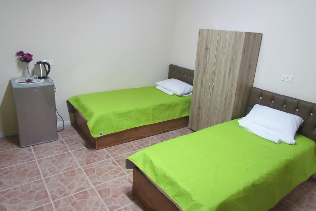 A room with two single beds, a wardrobe, a small table with a kettle and flowers, and a tiled floor.