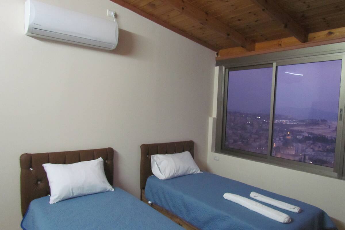 A bedroom with two beds, a window with a city view, and an air conditioning unit.