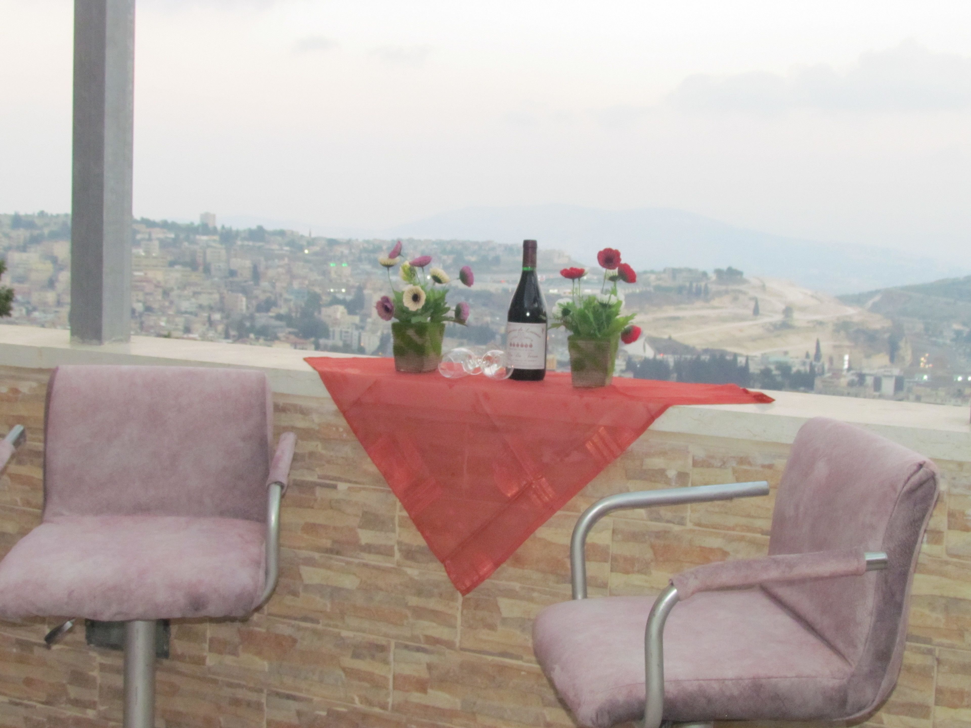Une table recouverte d’une nappe rouge, deux chaises, une bouteille de vin et de petites plantes en pot.