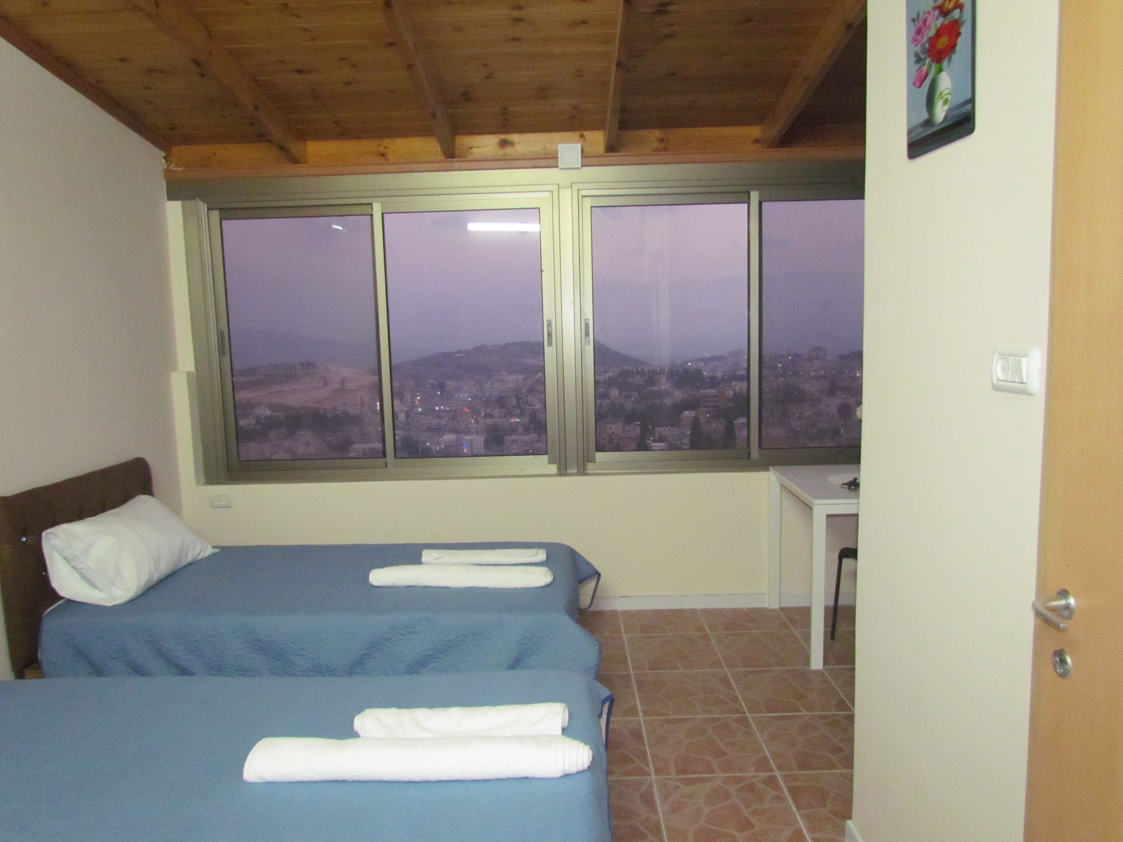 A bedroom with two beds, a desk, and a large window with a city view.