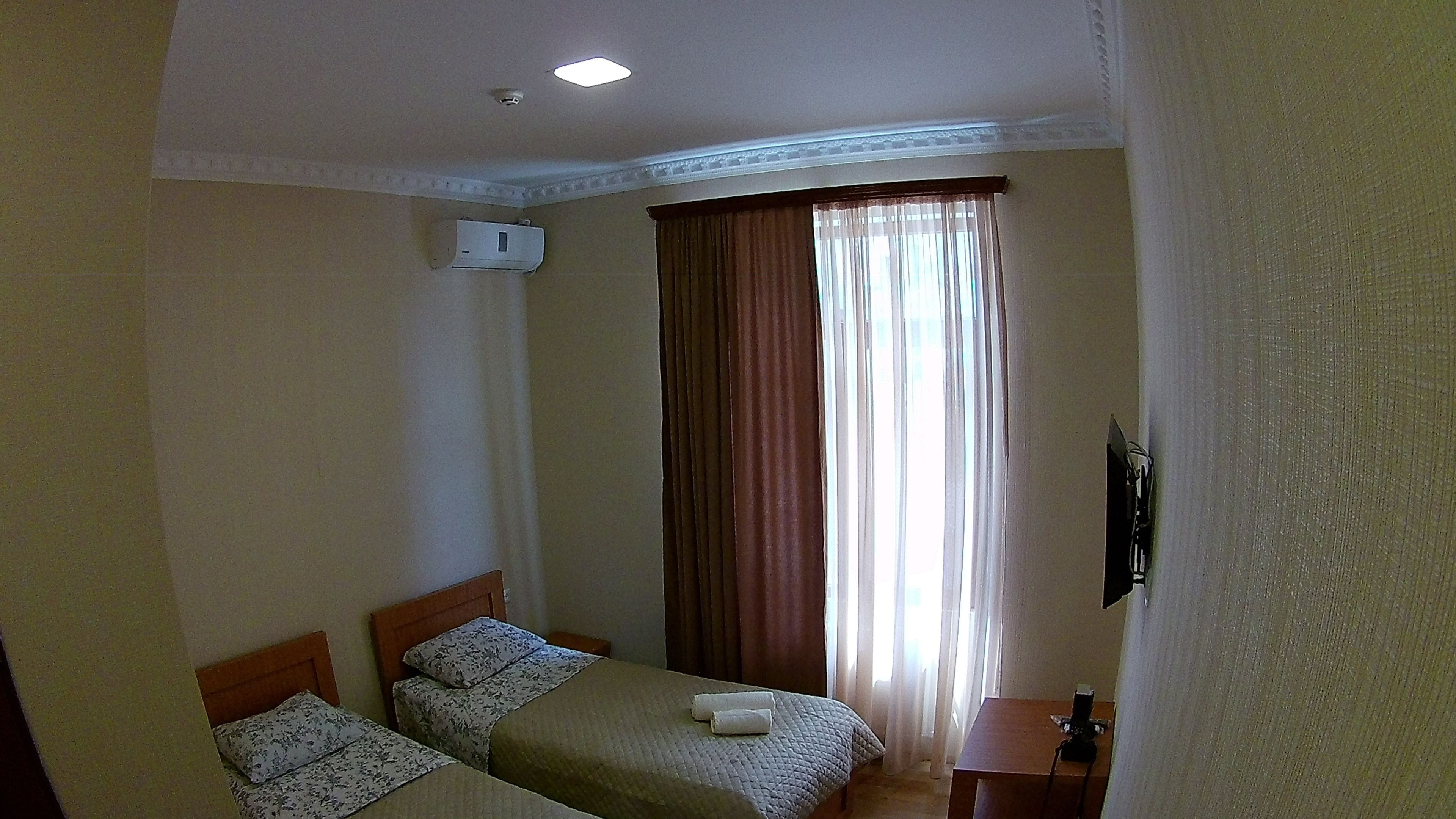 A hotel room with two single beds, a small table, a TV mounted on the wall, and a window with curtains.