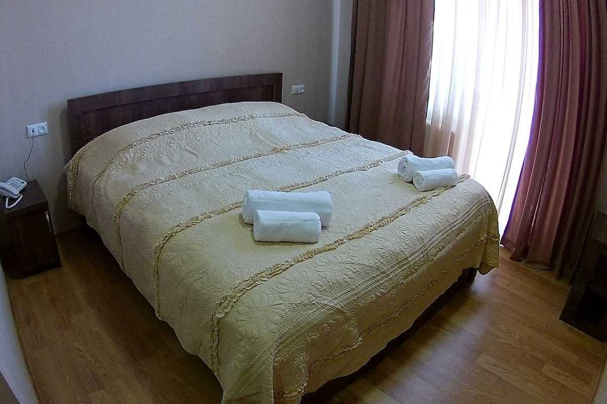 Ein ordentlich bezogenes Bett mit einer beigen Decke und zwei aufgerollten Handtüchern darauf.