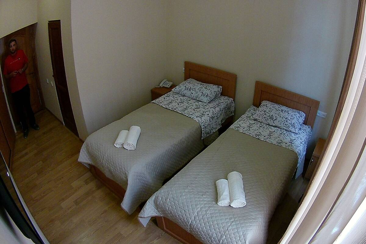 Ein Zimmer mit zwei Betten, jeweils mit einem Kopfkissen und einem zusammengerollten Handtuch. Es gibt ein hölzernes Kopfteil, einen Nachttisch mit Telefon und eine Tür zu einem weiteren Raum.